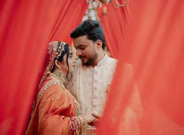 A couple in traditional Indian wedding attire posing behind red fabric curtains.