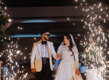 A bride and groom in a white tuxedo and sequined gown walk through indoor wedding sparklers.
