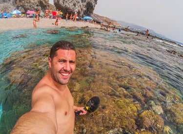 A man smiling for a selfie while snorkeling in the clear blue water of a Mediterranean beach.