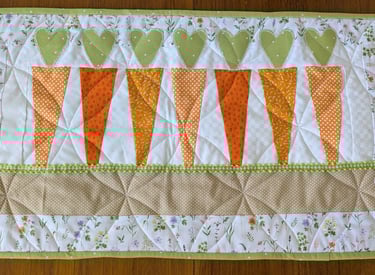 Quilted table runner with heart-topped carrots, floral border, and spring-themed stitching details.
