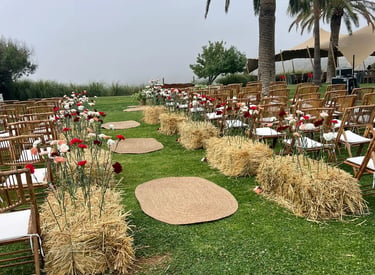 Outdoor wedding ceremony seating with wooden chairs, jute rugs, and colorful carnations growing from hay bales.