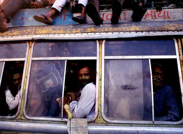 A side view of a heavily crowded yellow bus with passengers leaning out of windows and sitting on the roof.