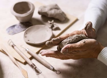 A pair of hands mold a piece of clay on a work surface, surrounded by various pottery tools and partially shaped clay objects