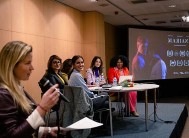 A panel of female speakers sits at a table during a film presentation for Mariana in an auditorium.