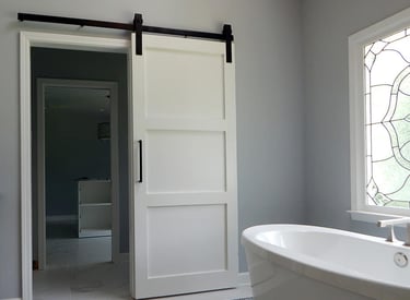 White sliding barn door with black hardware installed in a modern bathroom near a soaking tub.