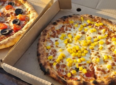 Two takeaway wood-fired pizzas in cardboard boxes with toppings of corn, olives, and cherry tomatoes.