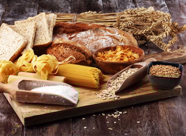 Variety of whole grain foods including bread, pasta, oats, and flour on a rustic wooden board.