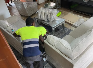 A professional mover in a yellow high-visibility vest with blue stripes bends over to wrap a large b