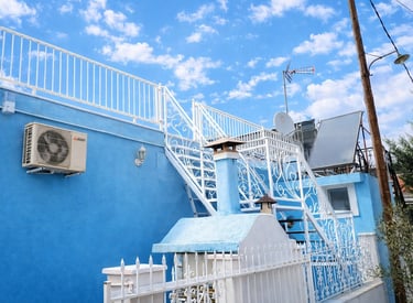 A blue Mediterranean house with white ornamental stairs, solar panels, and a blue sky with clouds.