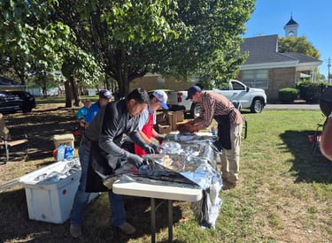 Volunteers at a community outdoor BBQ event preparing food on tables covered in aluminum foil.