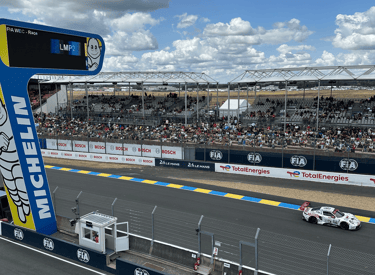 A white Porsche race car speeds past the grandstands at the 24 Hours of Le Mans circuit under a cloudy sky.