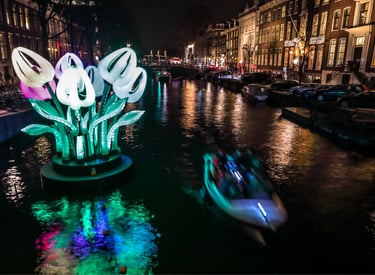 Illuminated giant tulip sculpture at the Amsterdam Light Festival on a canal with a passing tour boat.