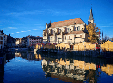 European Christmas market stalls reflecting in the blue river water by a historic stone church.