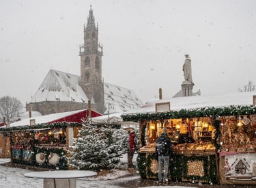 Snowy Bolzano Christmas market with wooden stalls and the Gothic cathedral spire in the background.