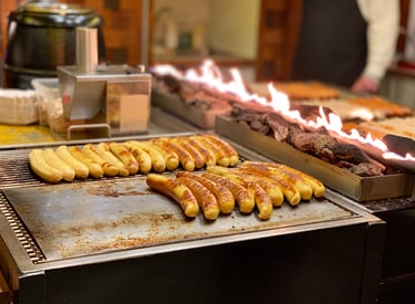 German bratwurst sausages grilling on a flat top stove next to an open charcoal flame at a food stall.