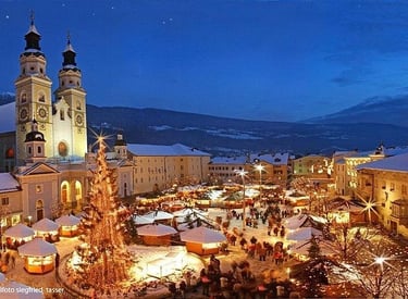A glowing Christmas market at night in Brixen, Italy, featuring a large lit tree and the Cathedral of Bressanone.