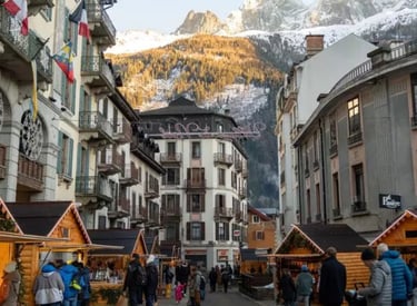Crowded Christmas market in Chamonix, France, with wooden stalls and snow-capped Alpine mountains in the background.