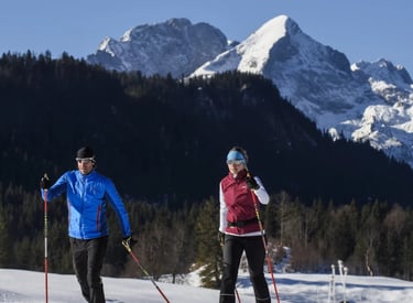 A man and woman cross-country skiing on a snowy trail with a scenic mountain range background.