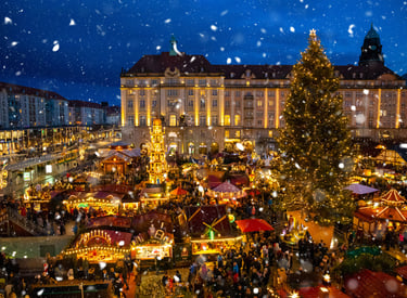 Festive Christmas market in Dresden at night with falling snow, glowing stalls, and a large decorated tree.