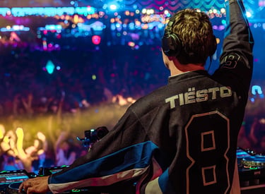 DJ Tiësto performing a live set on stage at an EDM music festival with a large cheering crowd and lights.