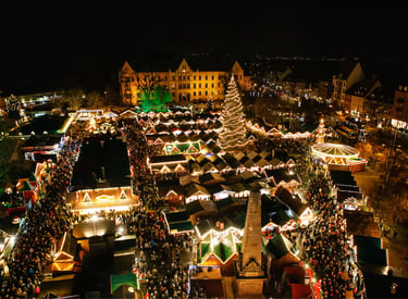 Aerial view of a festive German Christmas market at night with glowing stalls and a tall lit tree.