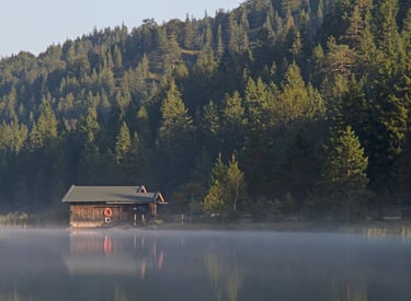 A rustic wooden boathouse on a misty mountain lake surrounded by a dense evergreen forest.