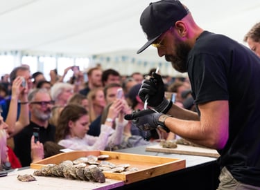 A professional chef shucking fresh oysters for a large crowd at a seafood festival demonstration.