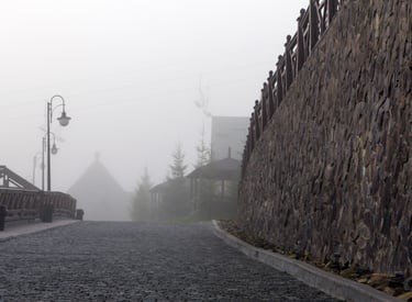 Foggy cobblestone road with a stone wall and street lamps in a misty mountain resort village.