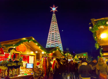 Crowds walking past illuminated wooden stalls and a giant crystal Christmas tree at an outdoor holiday market.
