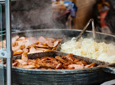 Steaming hot grilled bratwurst and sliced onions cooking in a large metal pan at a street food market.