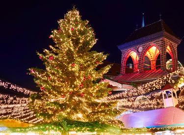 A glowing Christmas tree and festive market stalls decorated with holiday lights at night.
