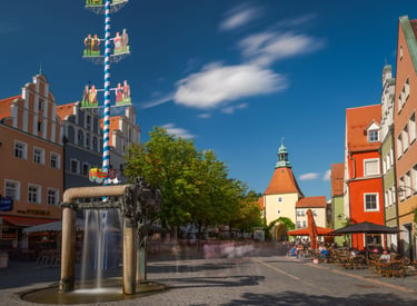 A sunny German town square featuring a traditional blue and white Maibaum maypole and historic architecture.