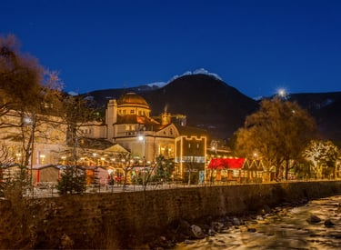 Illuminated Christmas market stalls in Merano, Italy at night with snowy mountains in the background.