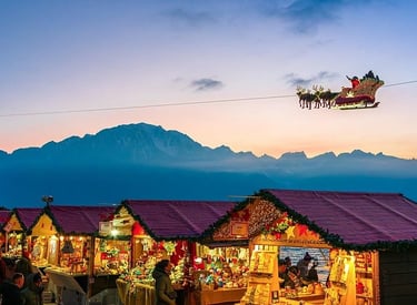 Christmas market stalls at dusk with Santa's sleigh flying over snowy mountains.