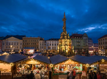 A bustling European Christmas market at dusk featuring the Holy Trinity Column in Olomouc, Czech Republic.