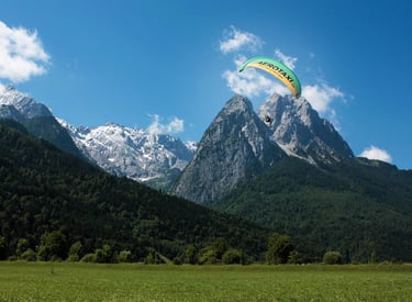 A green paraglider soaring over a lush valley and the snow-capped Julian Alps in Slovenia.