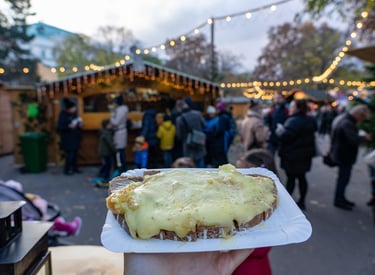A person holding melted raclette cheese on bread at a crowded outdoor European Christmas market.