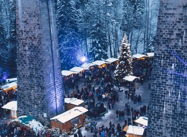 Crowded outdoor Christmas market in a snowy forest with wooden stalls and a tall decorated tree.