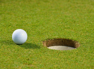 A white dimpled golf ball sits on the edge of a circular hole on a green grass golf course.