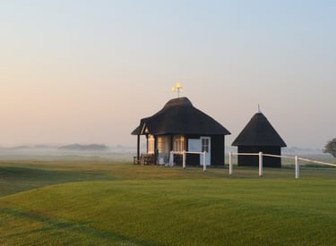 Small thatched-roof cottage and shed on a misty green golf course during sunrise.