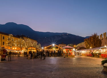 A festive European Christmas market at twilight with illuminated wooden stalls and mountains in the background.