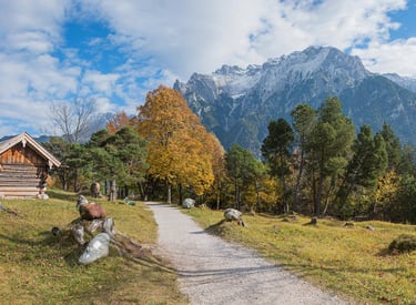 A rustic log cabin and gravel path in the Bavarian Alps with autumn trees and snow-capped peaks.