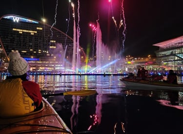 A person in a kayak watches a colorful firework and fountain show at night in Sydney's Darling Harbour.