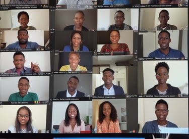 Three people are sitting around a conference table in a modern office. One person is wearing a red blazer and is speaking, while the other two listen and have open laptops in front of them. Papers with charts and graphs are scattered on the table. In the background, there are gray acoustic panels on the wall and a flip chart with colorful notes.