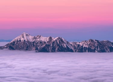 a snow covered mountain range poking above the clouds shrouded in pink purple evening sky