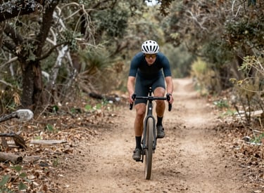 A cyclist riding along a peaceful trail winding through rolling hills and patches of forest under a clear blue sky.