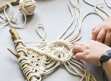Hands crafting a DIY macrame wall hanging using white cotton cord and a natural driftwood branch.