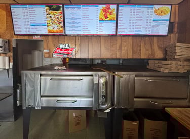 Professional pizza oven in a restaurant kitchen below digital menu boards displaying pizza and grinder prices.