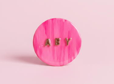 Pop socket para teléfono en tono rosa personalizado, con iniciales ABY en dorado.