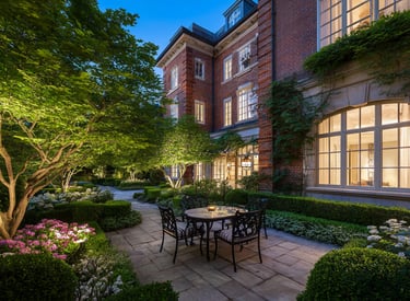 Luxury brick estate features a stone patio with a dining set and lush garden landscaping at dusk.